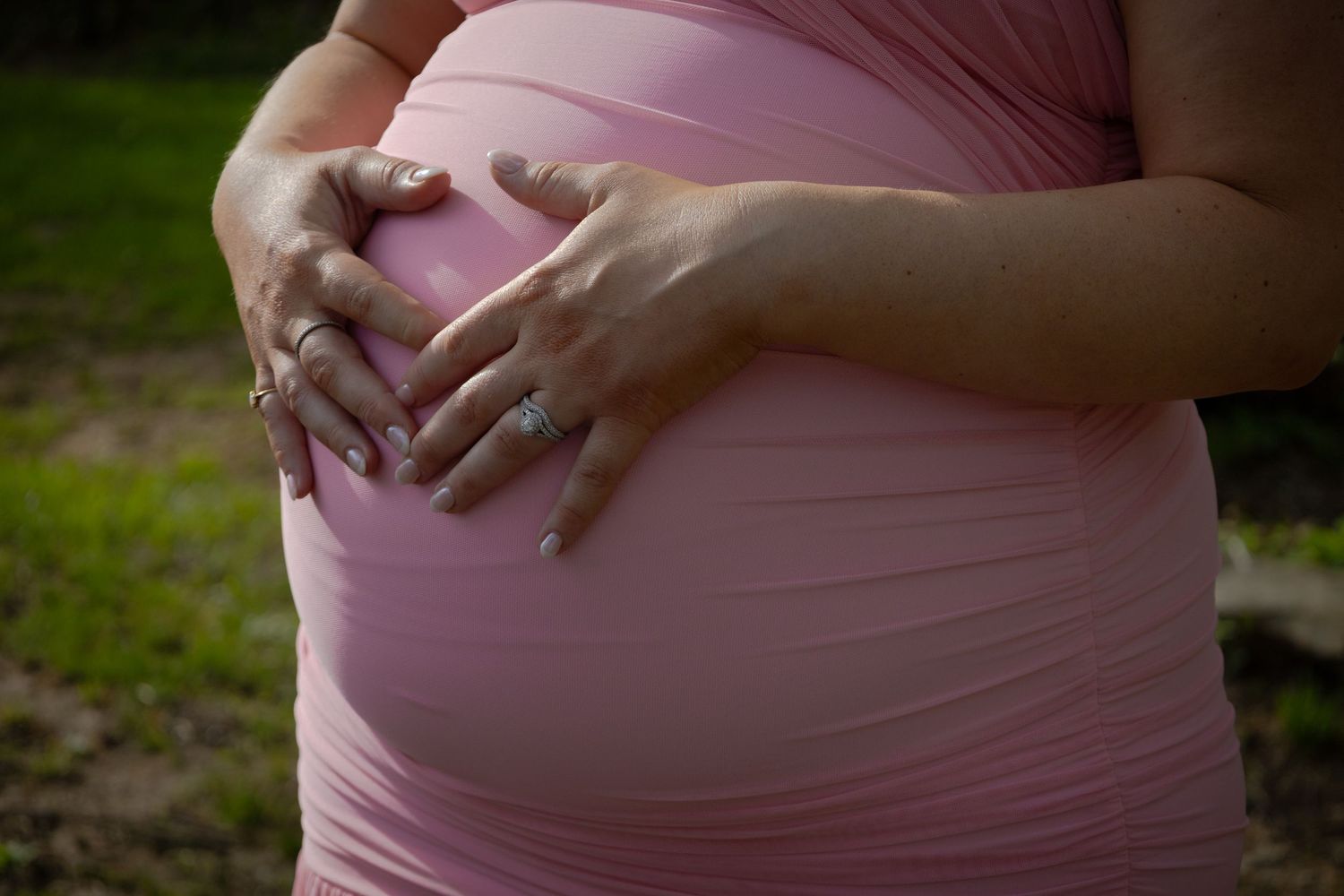Close-up view of hands cradling a pregnant belly wearing a pink maternity dress outdoors.