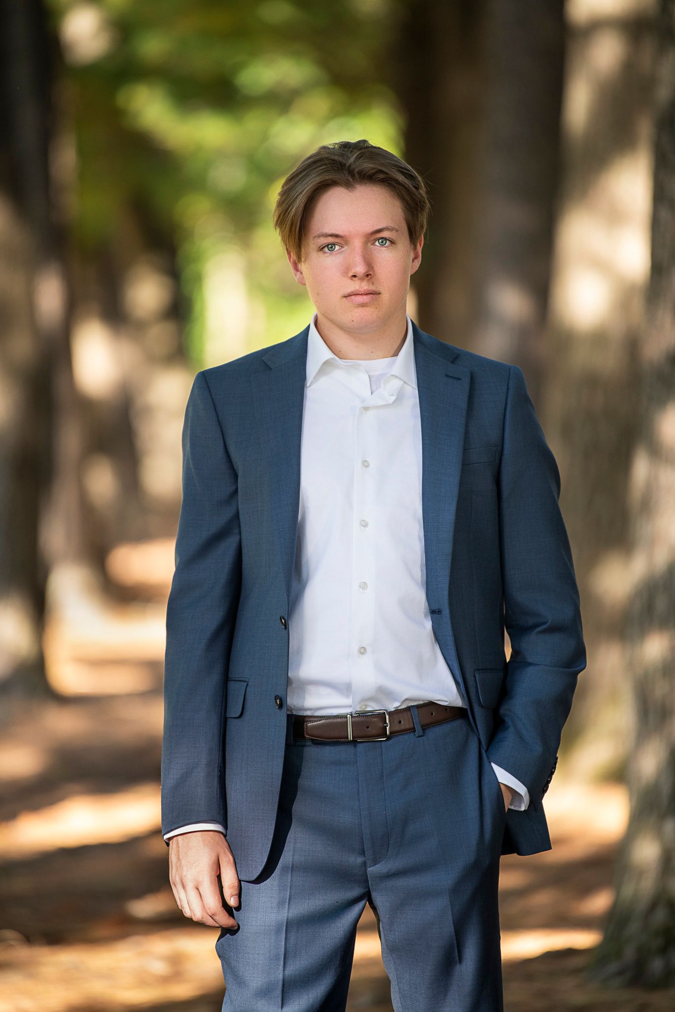 A person wearing a navy blue suit and white dress shirt poses outdoors in a wooded setting with soft natural lighting.