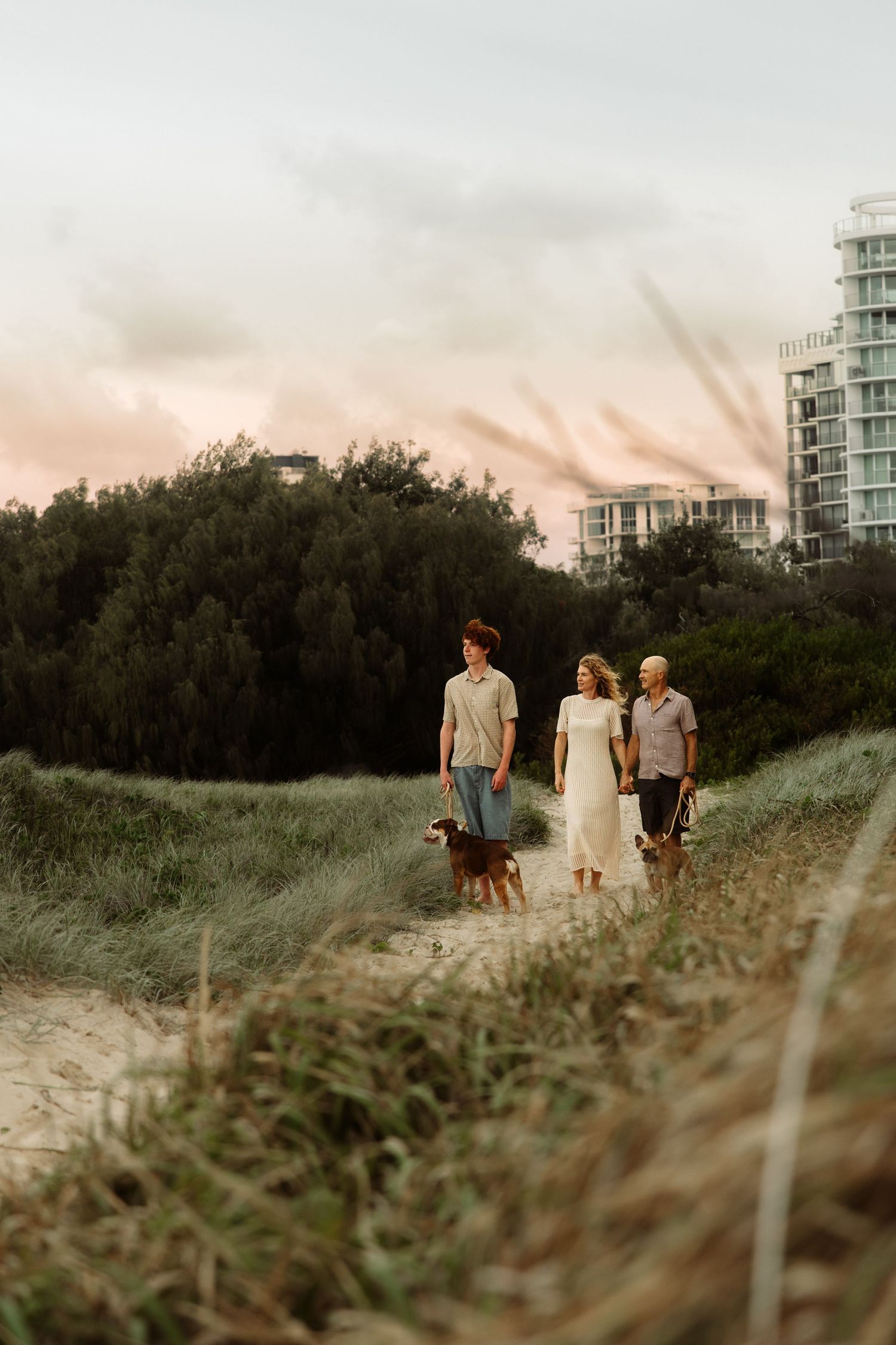 Three people and a dog walk along a grassy coastal path at sunset with modern buildings in the background.