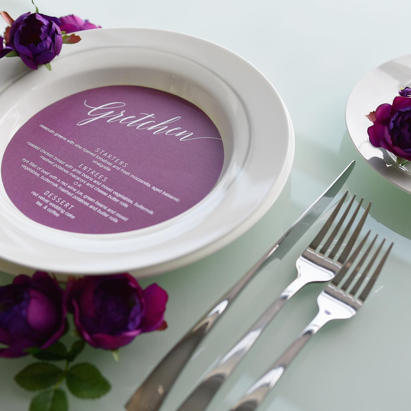 Round white plate with purple circular menu card and deep purple roses alongside silver cutlery on light surface.