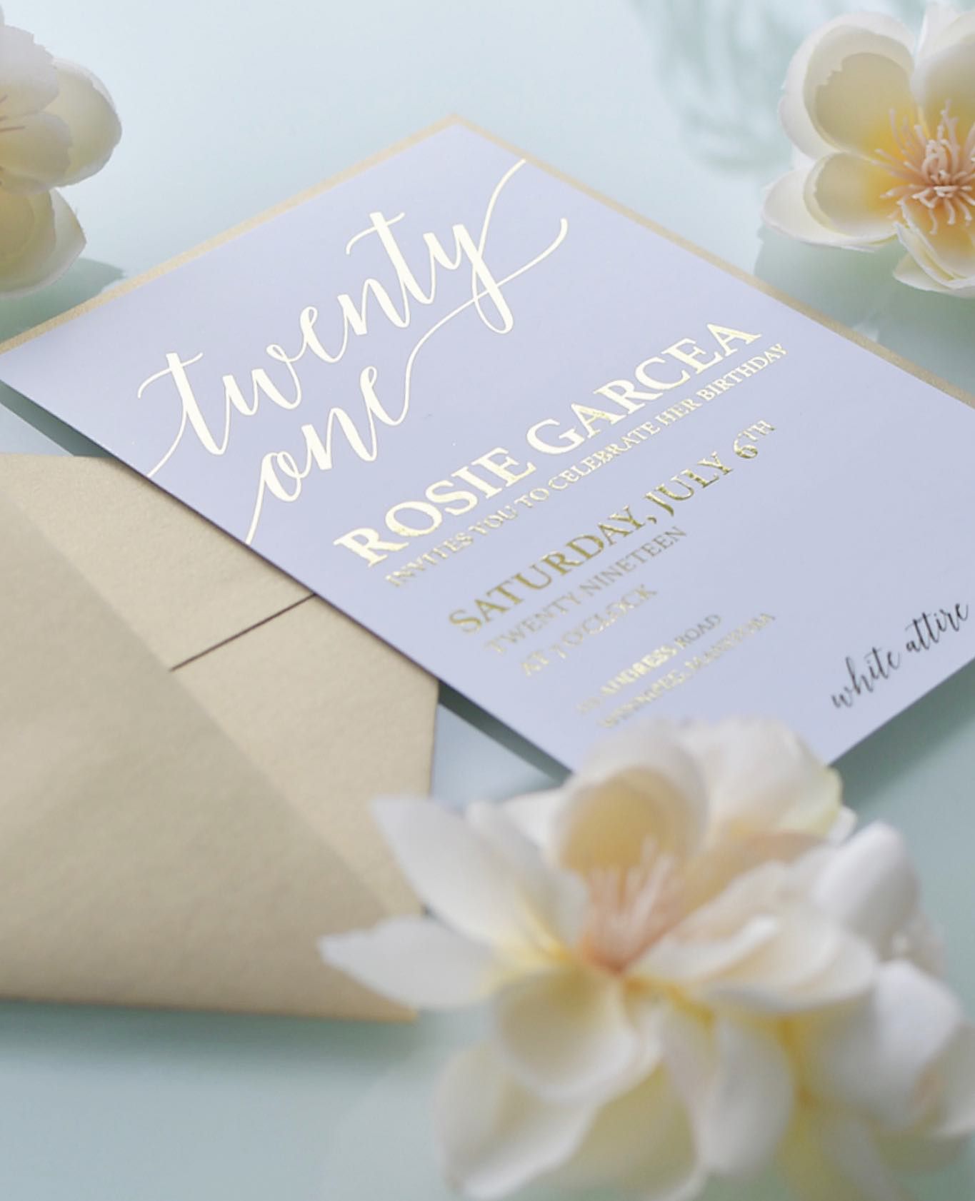 Wedding invitation in lavender tones with calligraphy text and delicate white plumeria flowers against soft lighting.