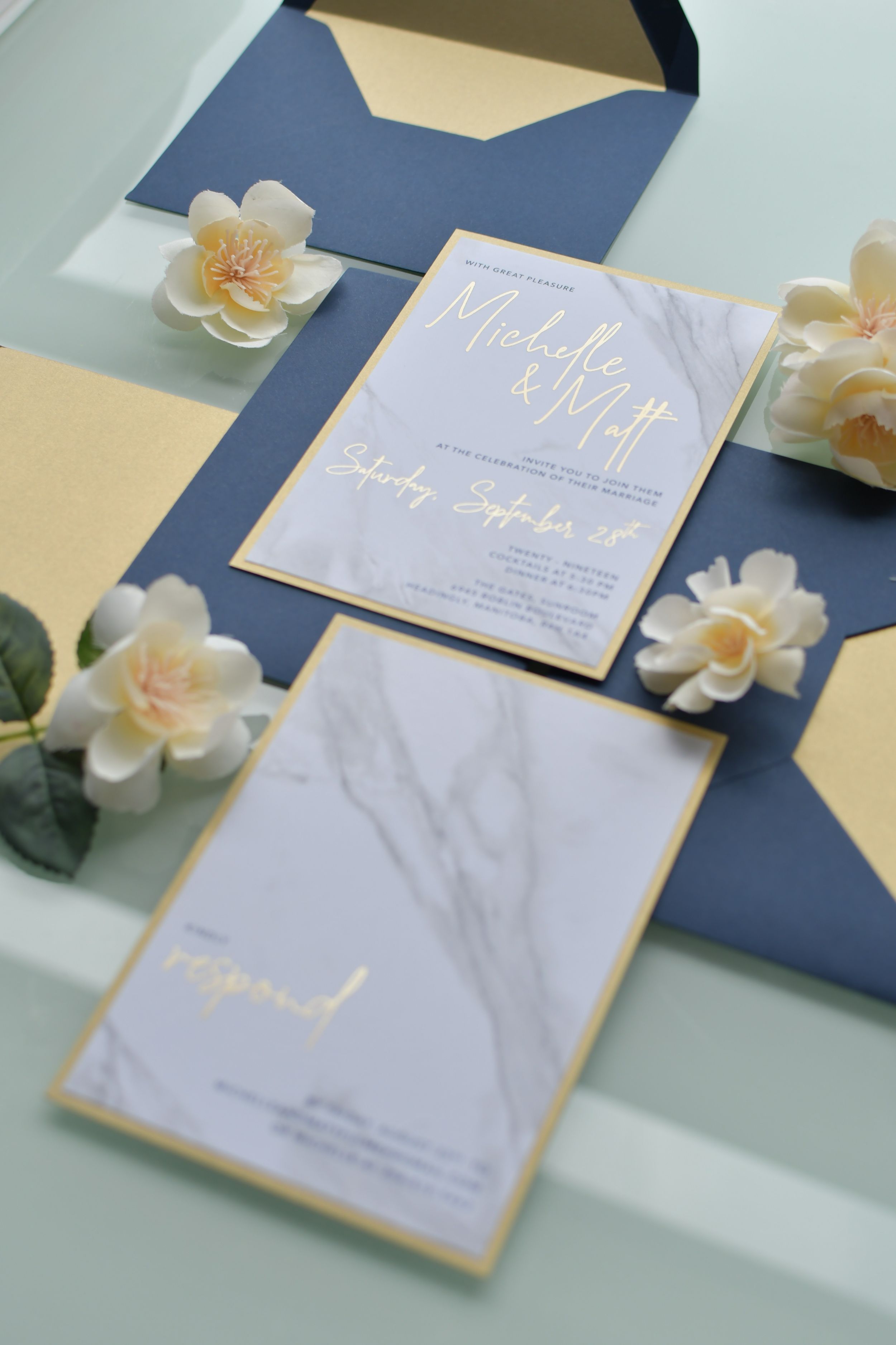 Elegant wedding invitation suite in navy blue and marble with gold accents and peach roses on a light blue surface.