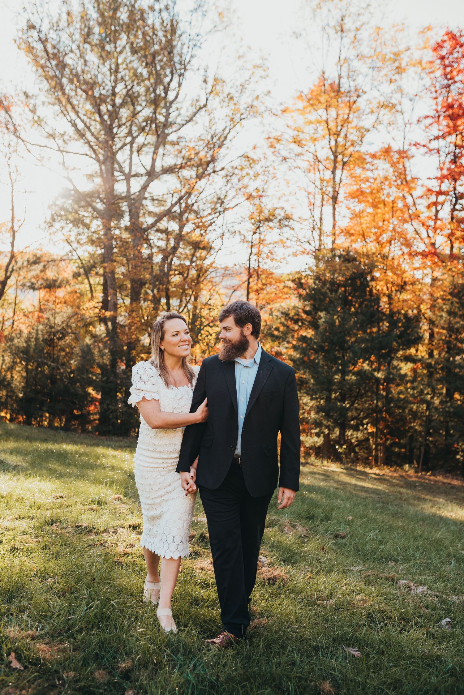 A couple walks together on a grassy hill with vibrant fall foliage in the background.