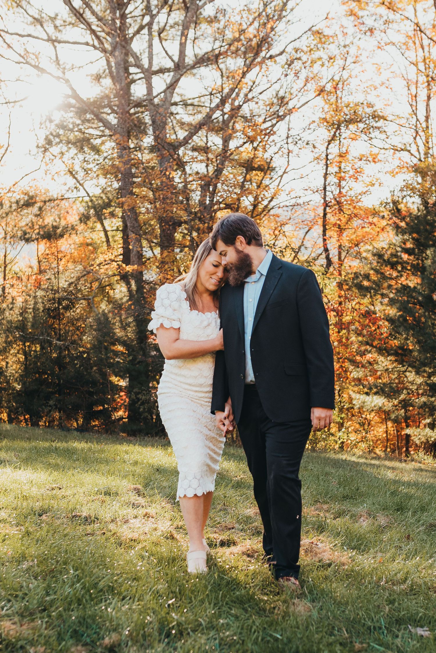 A couple shares a tender moment on a grassy hillside surrounded by autumn trees.