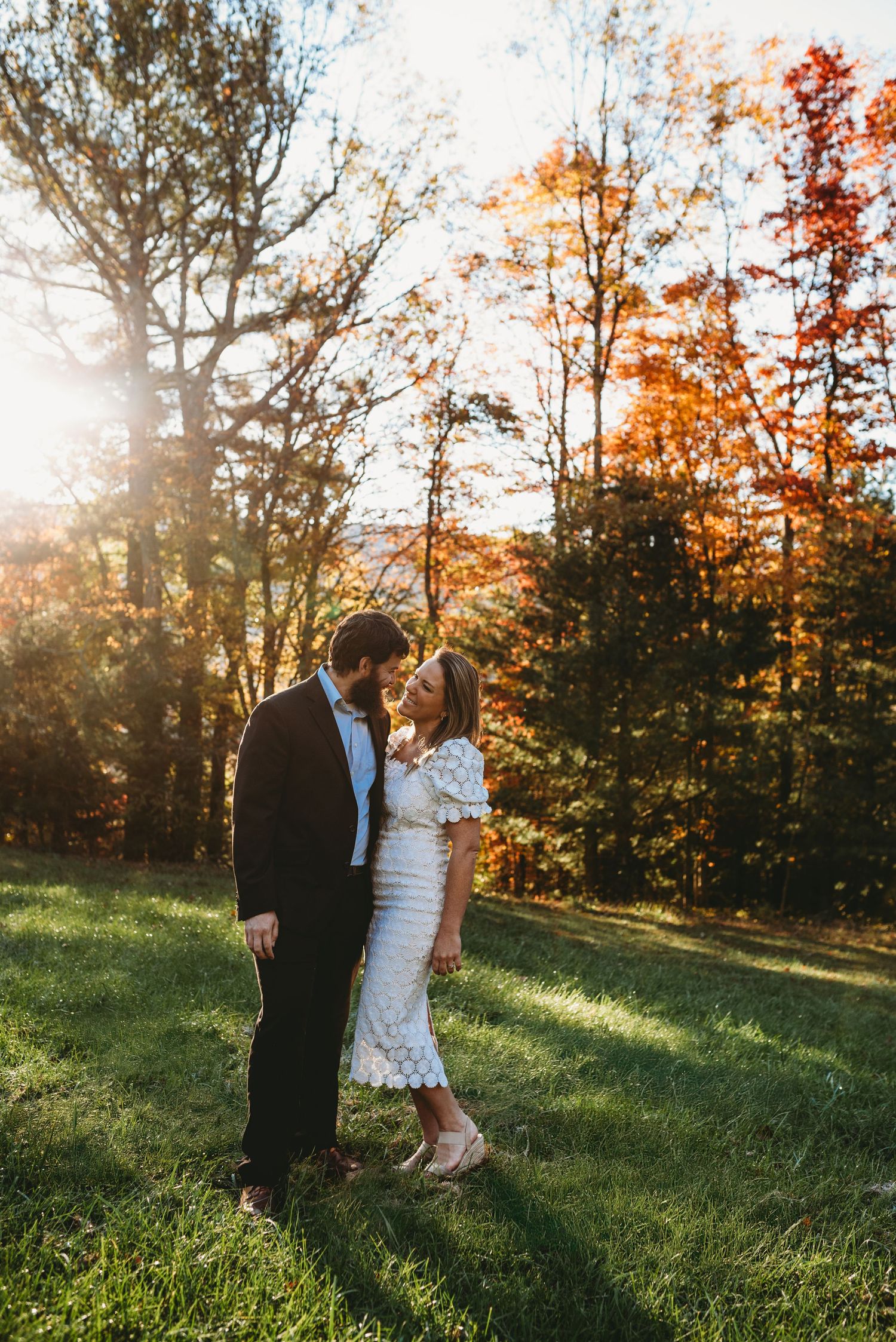 A romantic couple embraces in a sun-lit autumn field with colorful fall trees in the background.