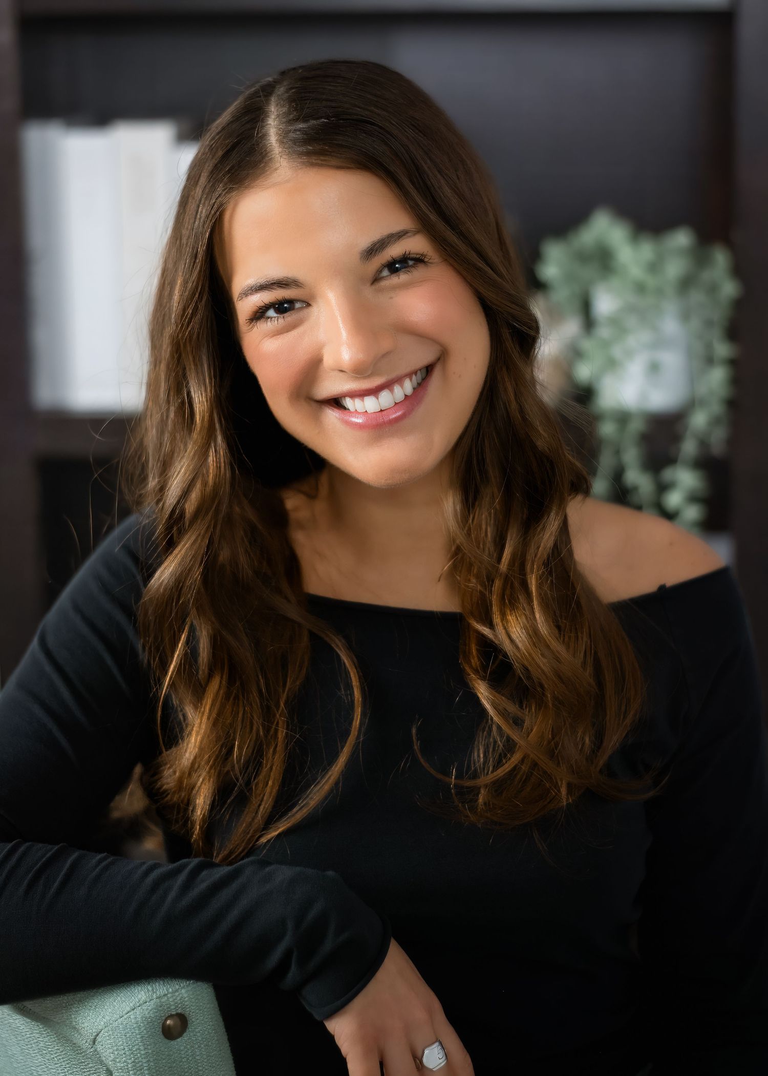 Professional headshot of someone wearing a black long sleeve top smiling warmly against a dark background with indoor plants.
