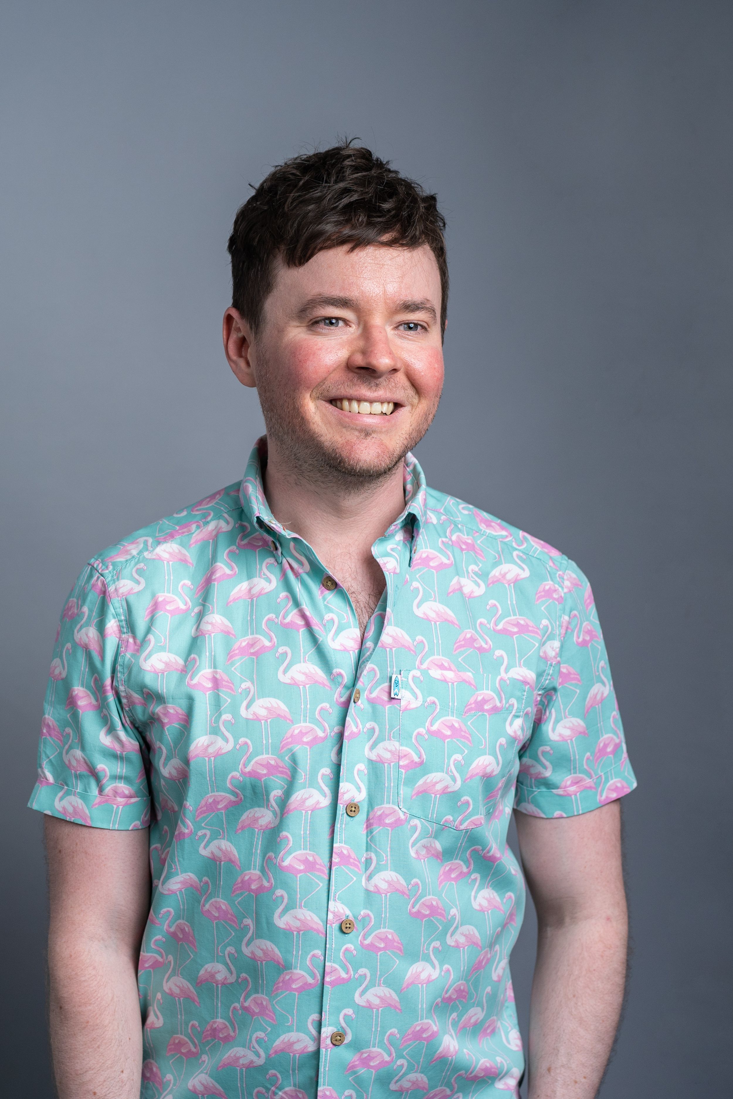A person wearing a turquoise short-sleeve button-up shirt with pink flamingo print stands against a gray background.
