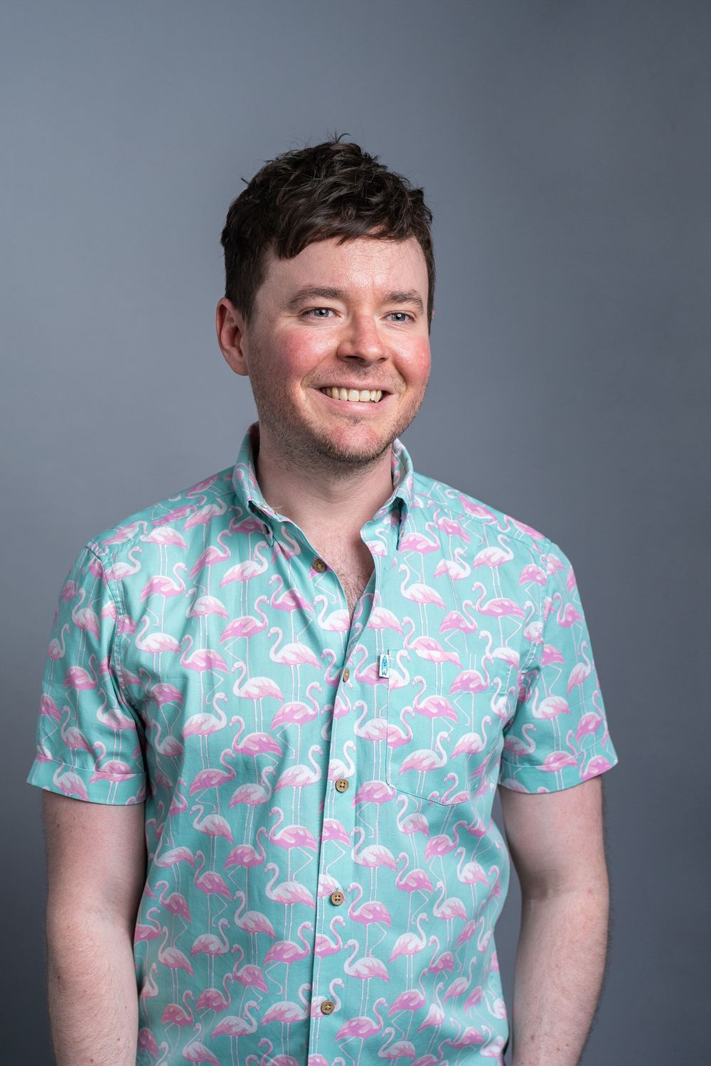 A person wearing a turquoise short-sleeve button-up shirt with pink flamingo print stands against a gray background.