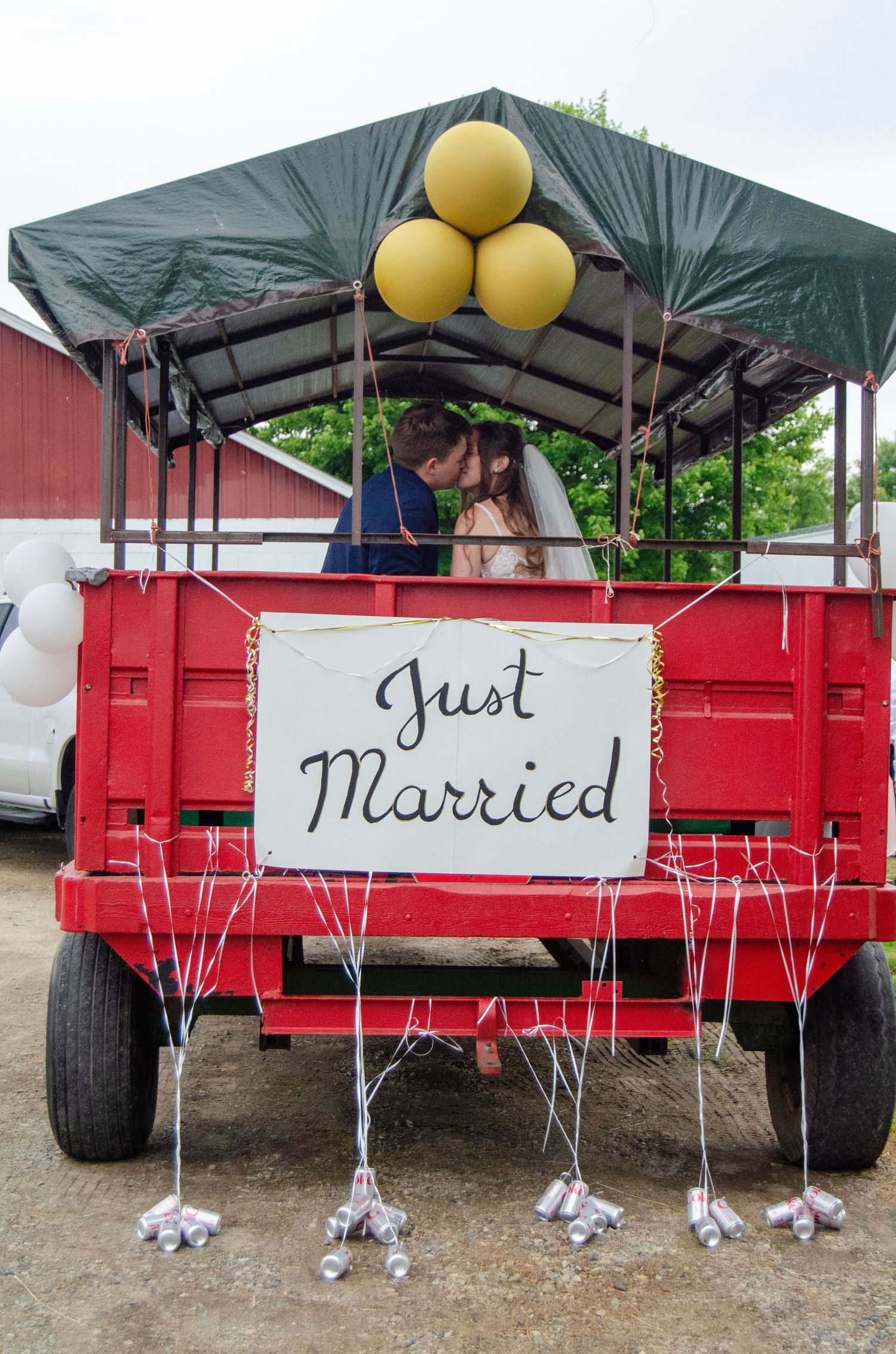 Red farm wagon decorated with 'Just Married' sign and white ribbons with shoes hanging underneath for rustic wedding decor.