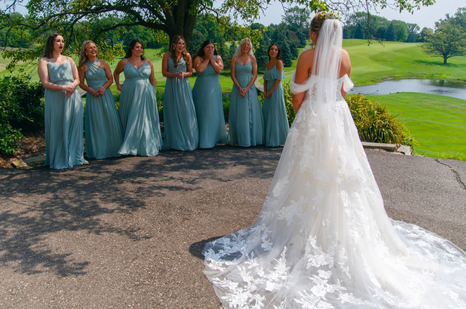 A wedding party in sea foam green bridesmaid dresses stands outdoors with a bride wearing a long lace train wedding gown.