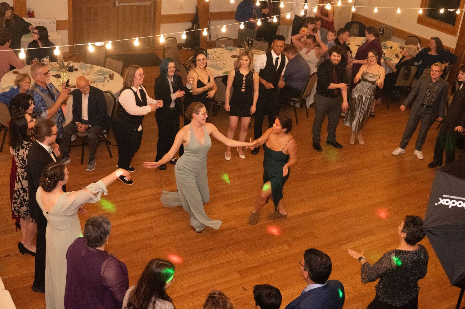 People dancing together in a large wooden-floored room with string lights overhead during a social dance event.