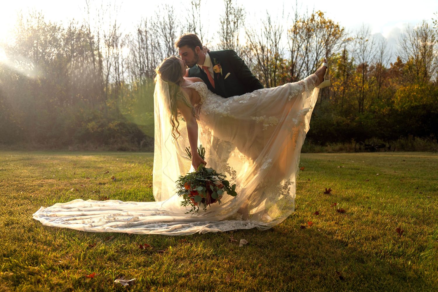 A couple in flowing wedding attire dances together in a sunlit meadow during golden hour.