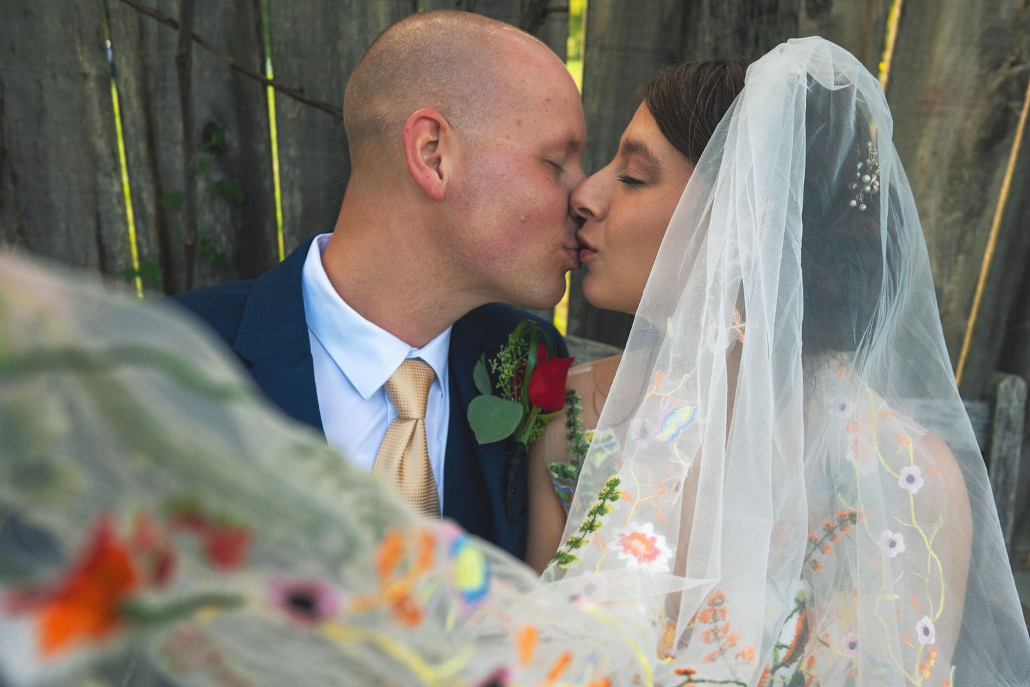 A romantic wedding kiss moment captured through a white veil with colorful flowers in the background.
