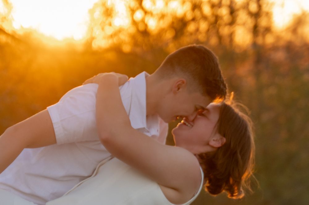 A romantic couple embraces during golden hour sunset with warm orange light glowing through trees in background.