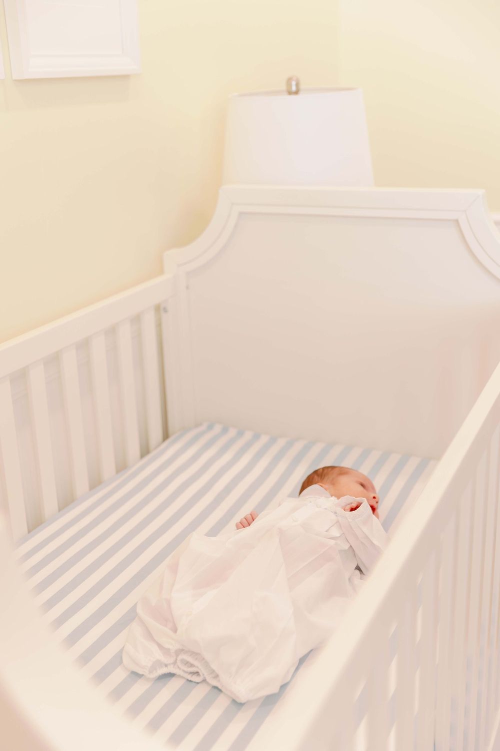 Sleeping newborn in white swaddling blanket rests peacefully in classic white crib with striped bedding.