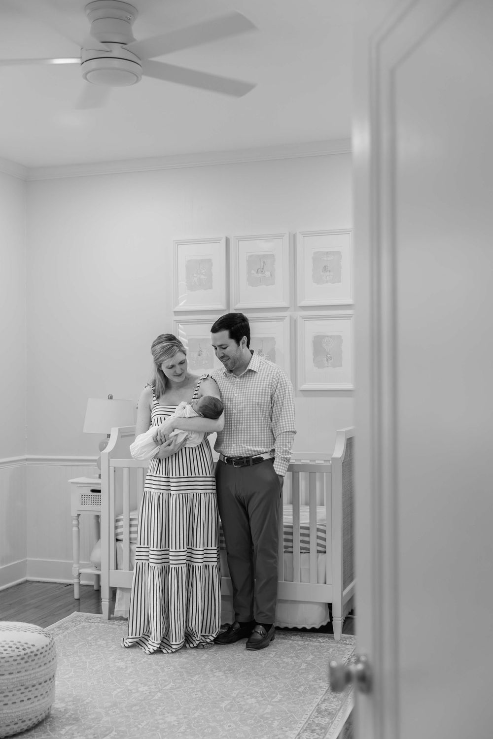 A couple stands together in a white nursery room with framed artwork and a ceiling fan.
