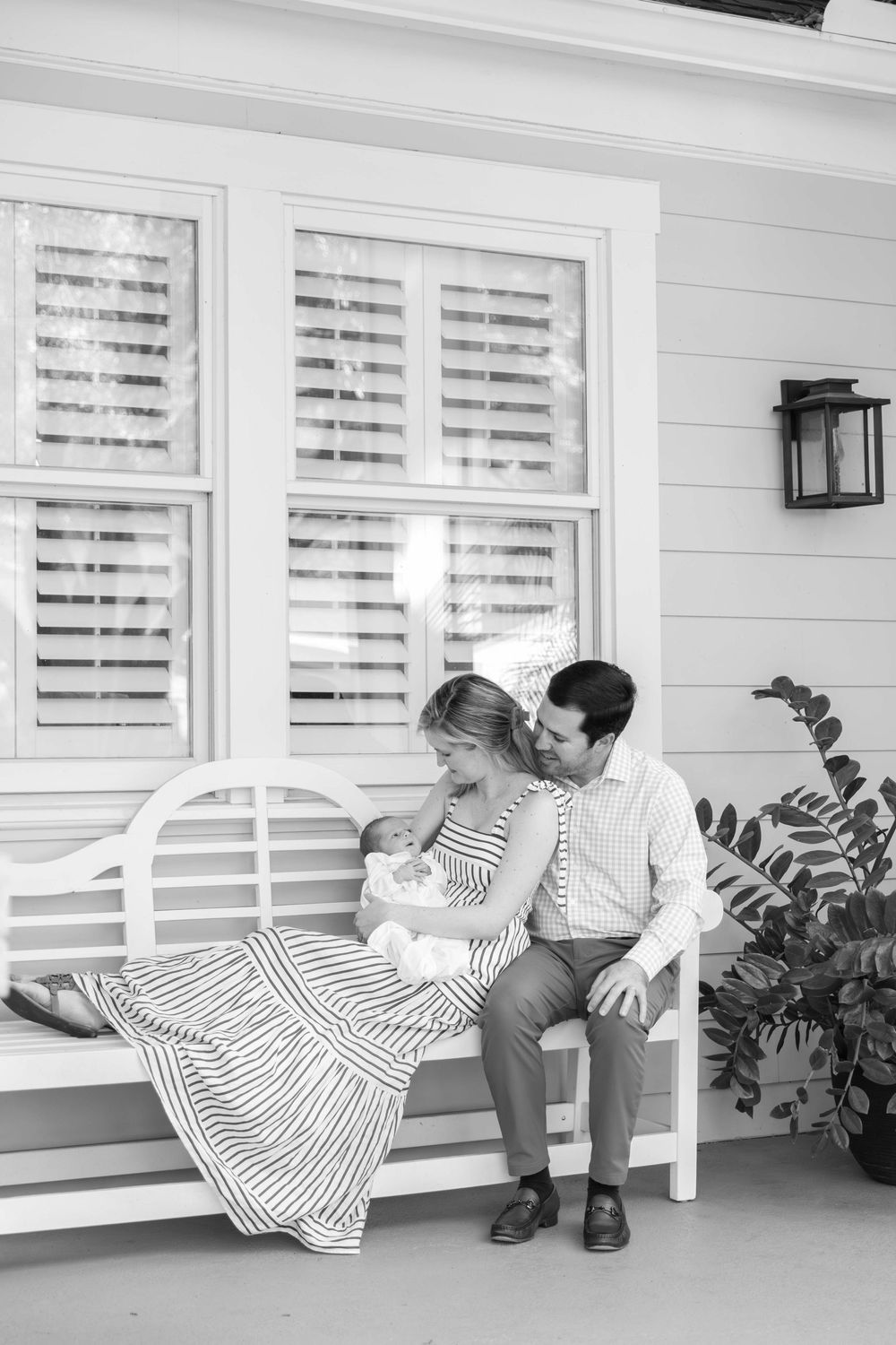 Family lounges together on white porch bench near shuttered windows and outdoor lantern in black and white photo.