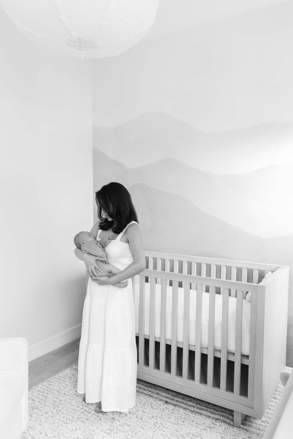 A person wearing white leans over a white wooden crib in a minimalist nursery setting.