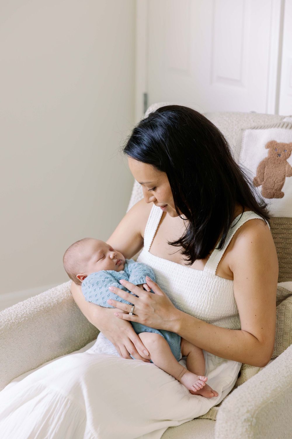 Mother in white tank top gently holds and looks down at infant in blue onesie while sitting in cozy armchair.