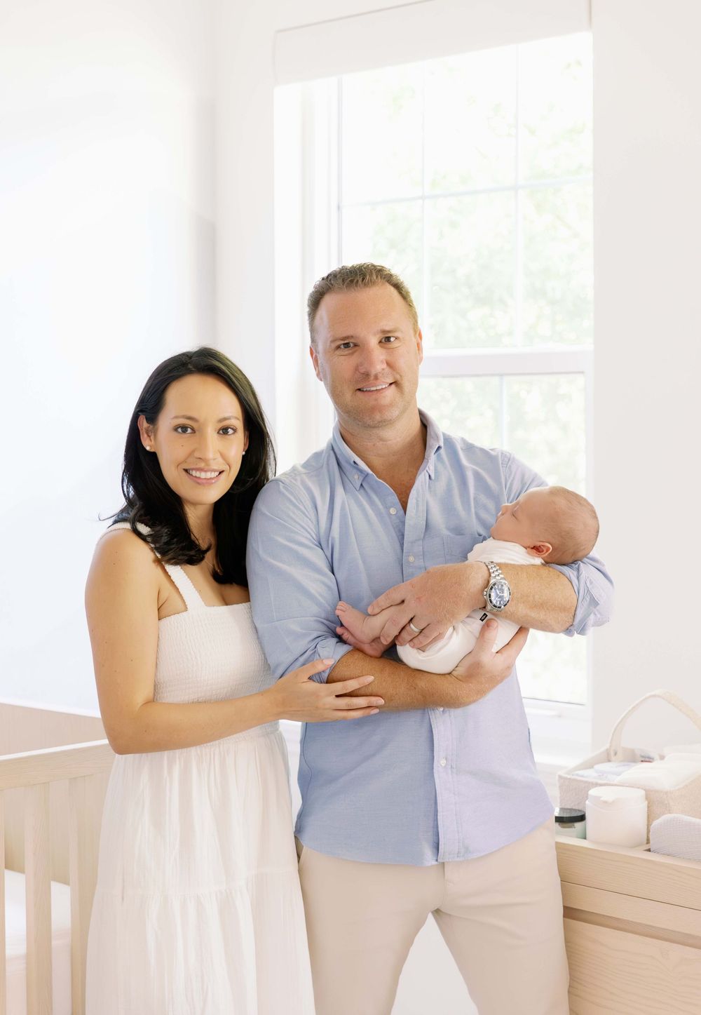 A couple stands together holding their newborn baby in a bright, airy nursery with white decor.