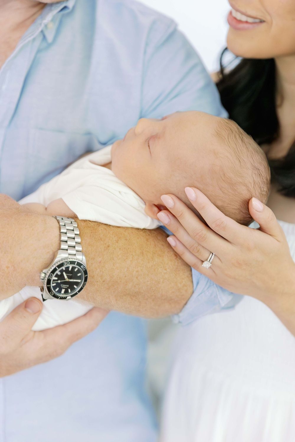Close up view of hands wearing a silver watch and wedding ring while cradling a newborn baby.