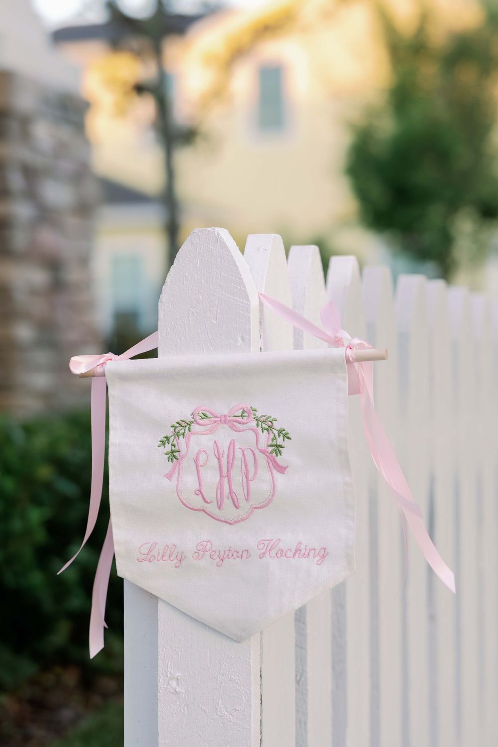 A white decorative banner with pink monogram and ribbons hangs on a picket fence post.