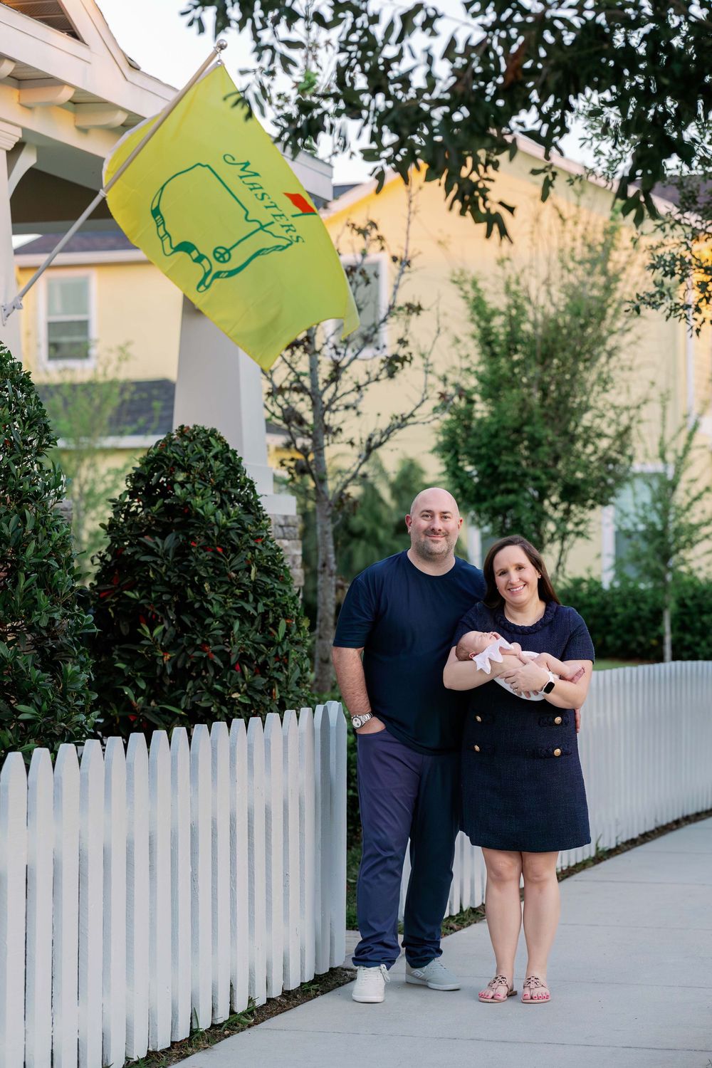 Family standing by white picket fence with Masters tournament flag displayed outside their home.