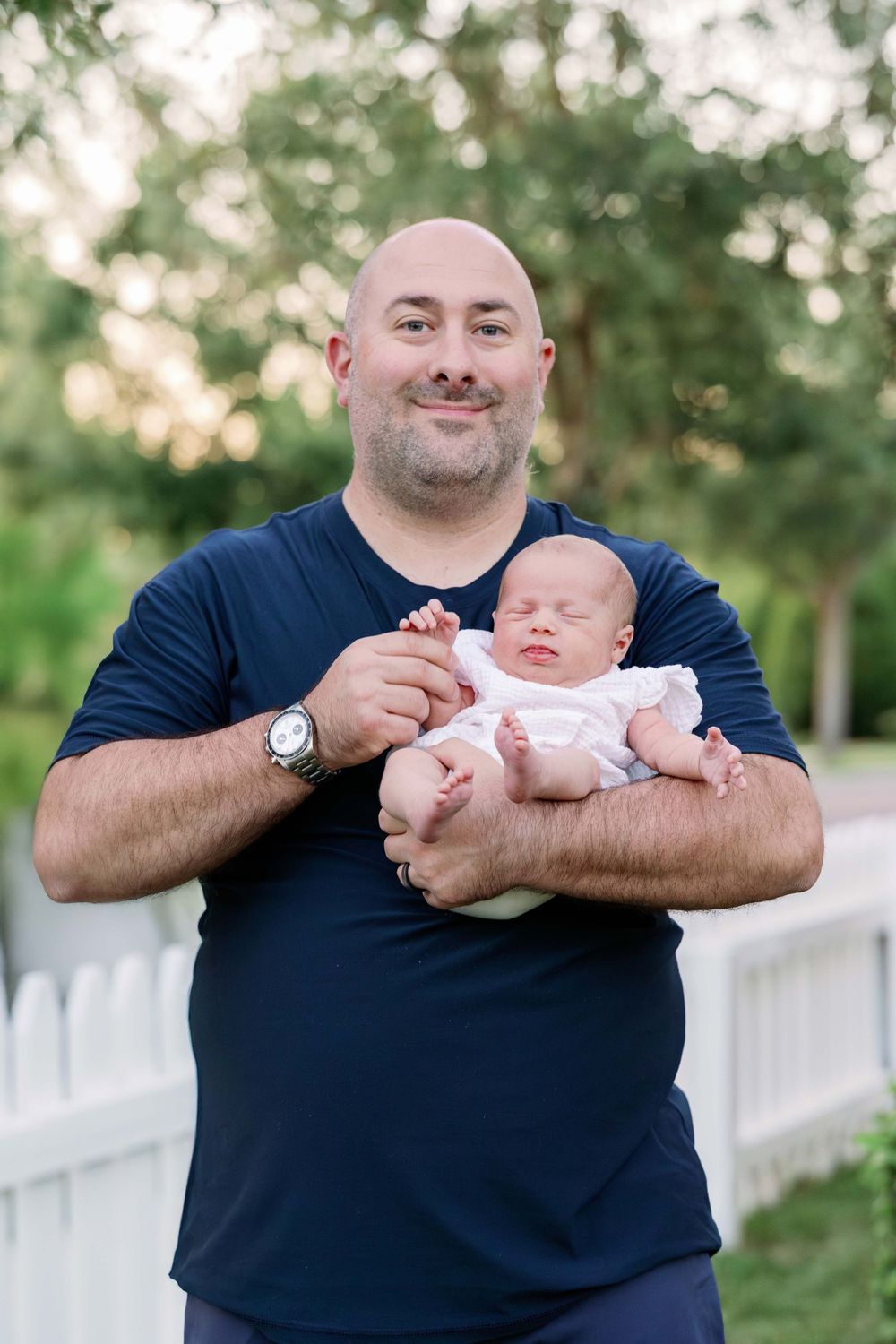 A parent in a navy shirt holds a sleeping newborn baby outdoors in front of a white picket fence and garden.