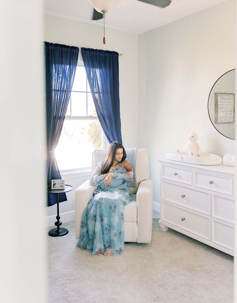 A nursery room features navy blue curtains, white furniture, and a cozy glider chair by a sunlit window.
