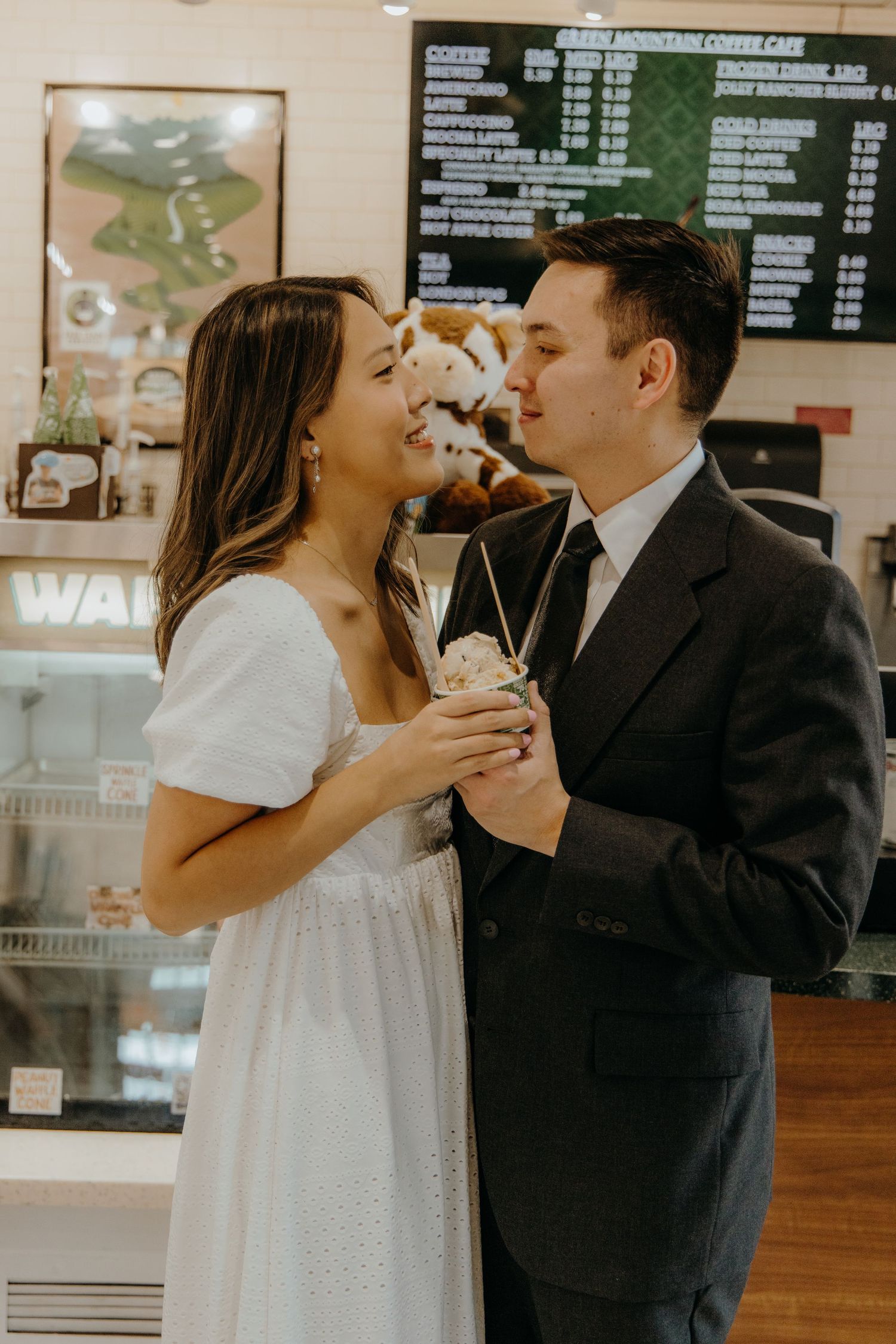 Couple sharing a milkshake at a coffee cafe, woman in white dress and man in dark suit smiling at each other.
