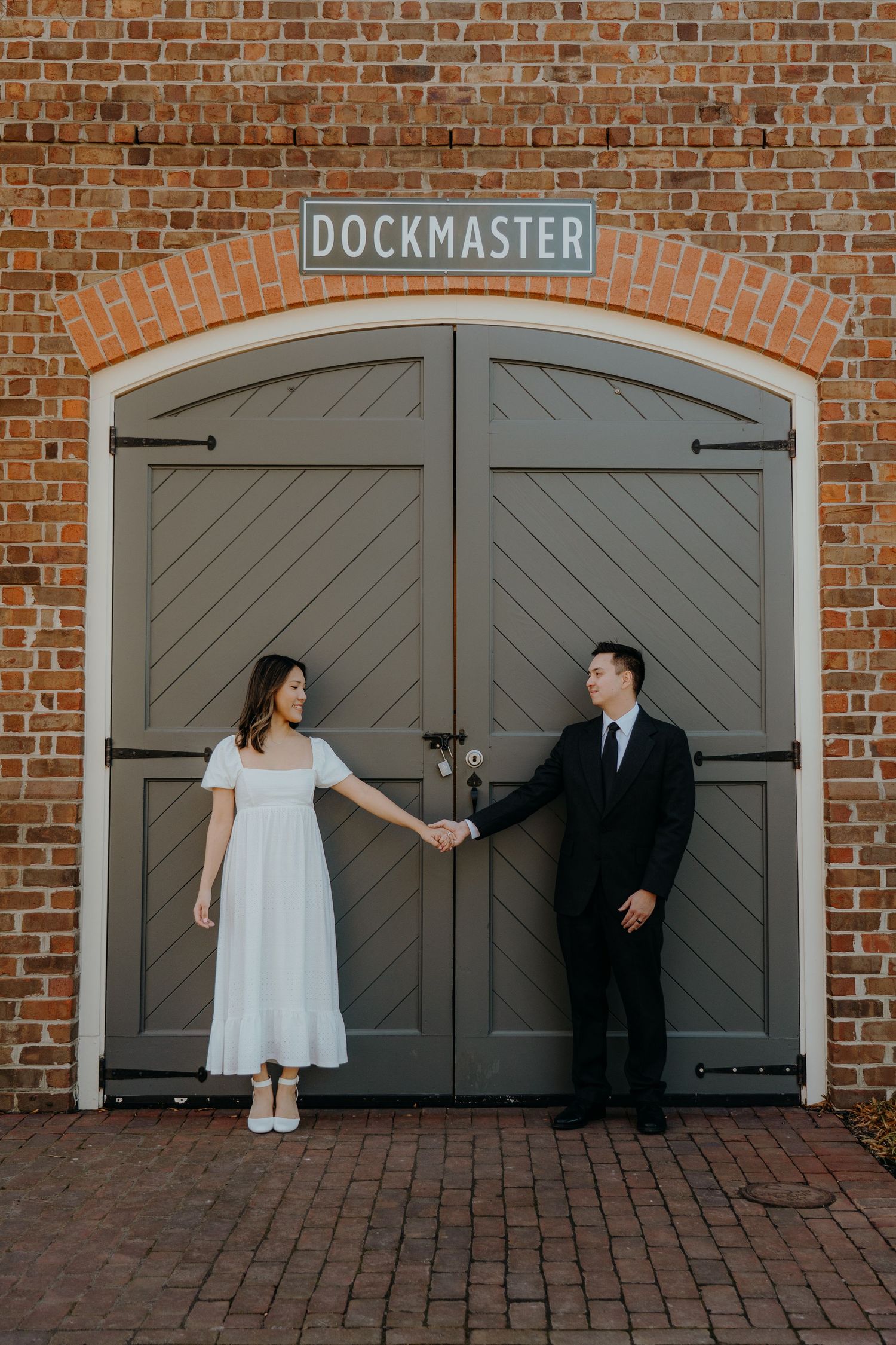 Couple holding hands in front of a gray arched Dockmaster door on a brick building, bride in white dress, groom in black suit.