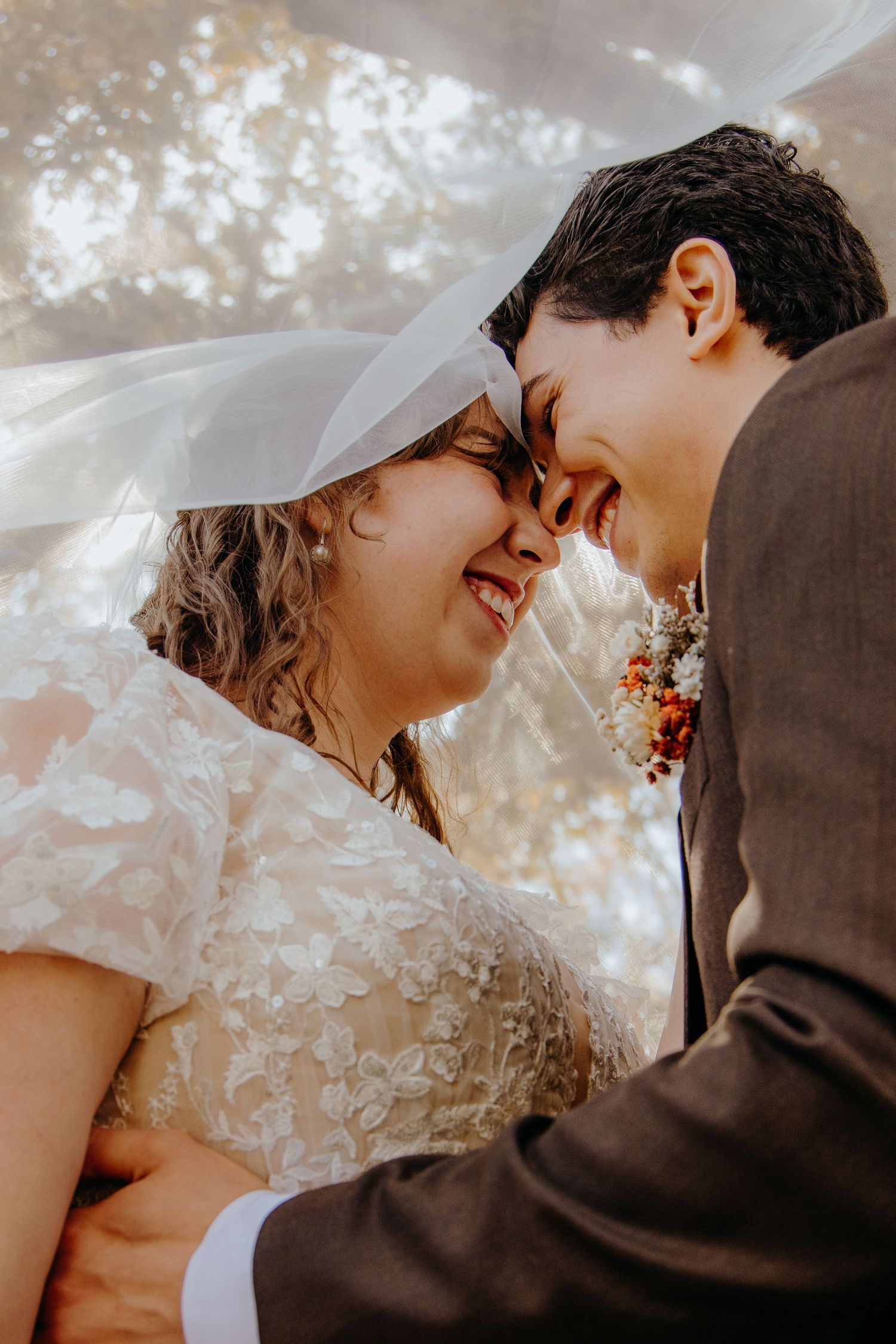 A joyful bride and groom share an intimate moment under a flowing veil, smiling and touching foreheads on their wedding day.