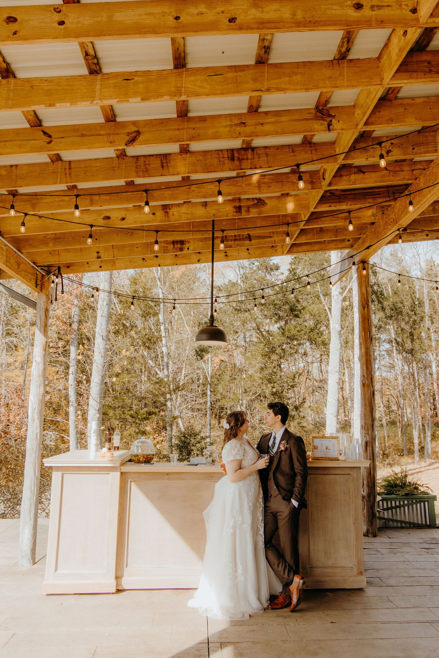 Bride and groom share a romantic moment at an outdoor bar under a wooden pergola with string lights and forest backdrop.