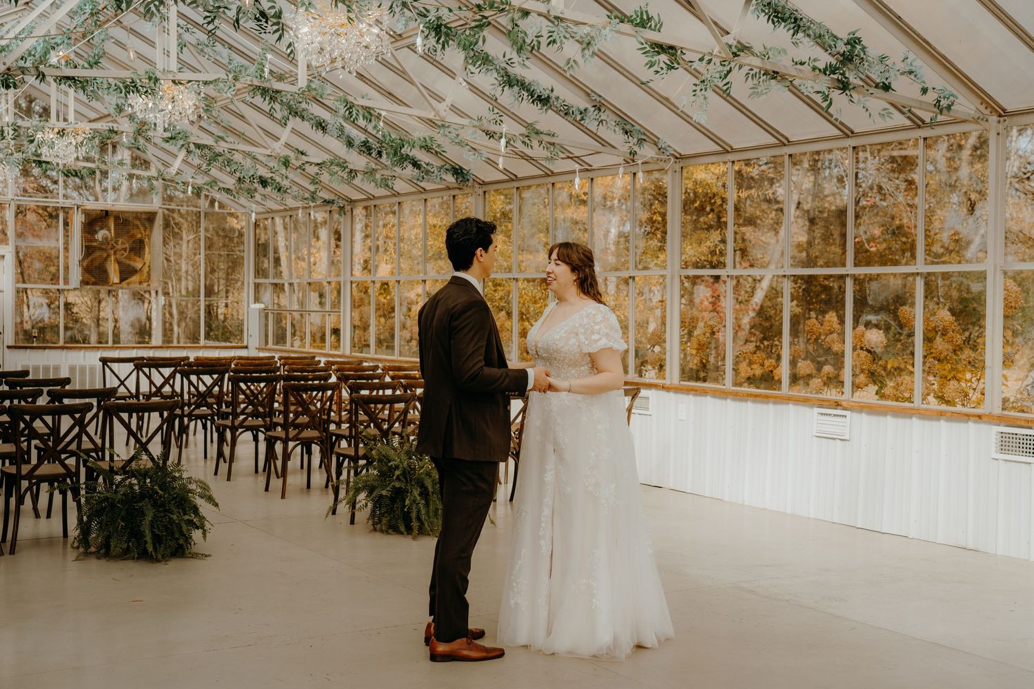 Bride and groom share a first look in an elegant glass greenhouse wedding venue with wooden chairs and greenery.