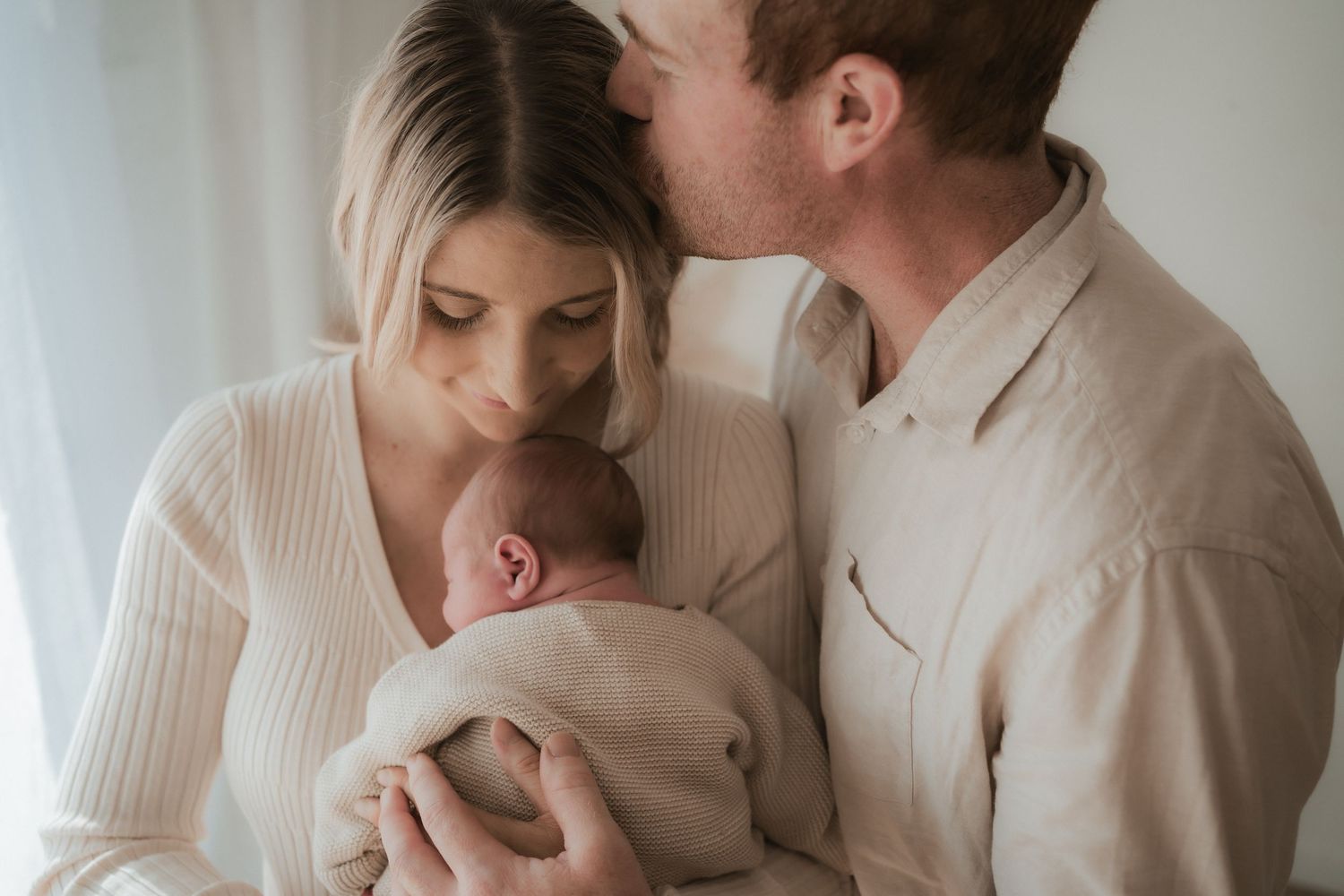 Parents in soft neutral clothing embrace their newborn baby while standing near window in intimate family moment.