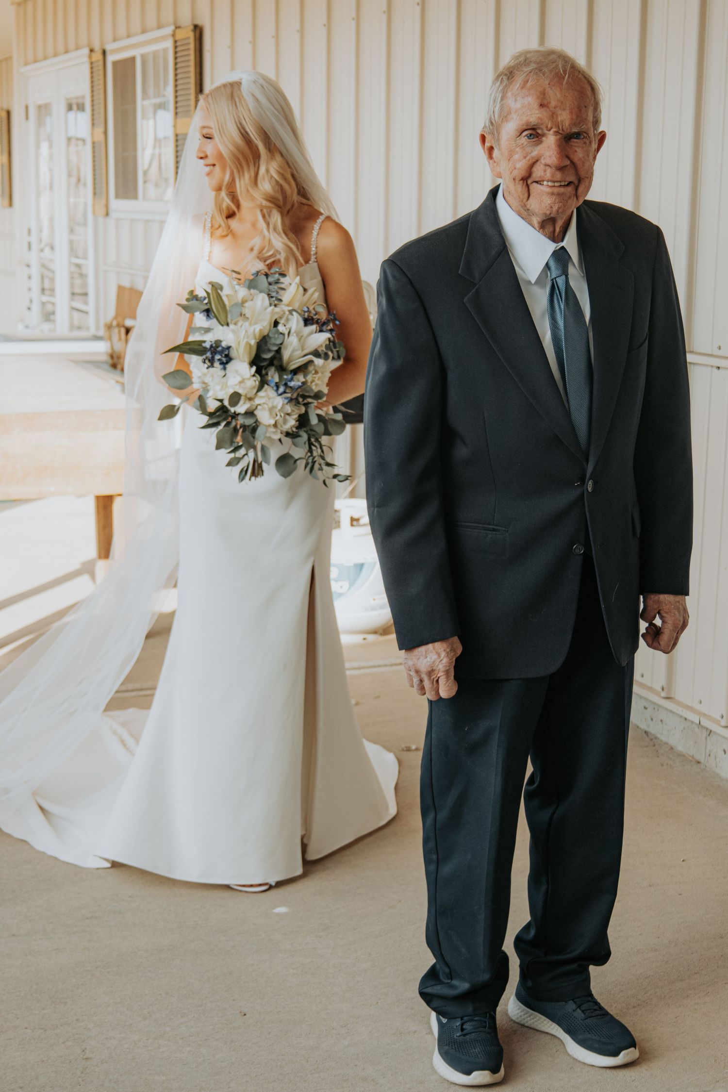 A bride in a white dress holding a floral bouquet stands next to an older gentleman in a dark suit at a wedding venue.