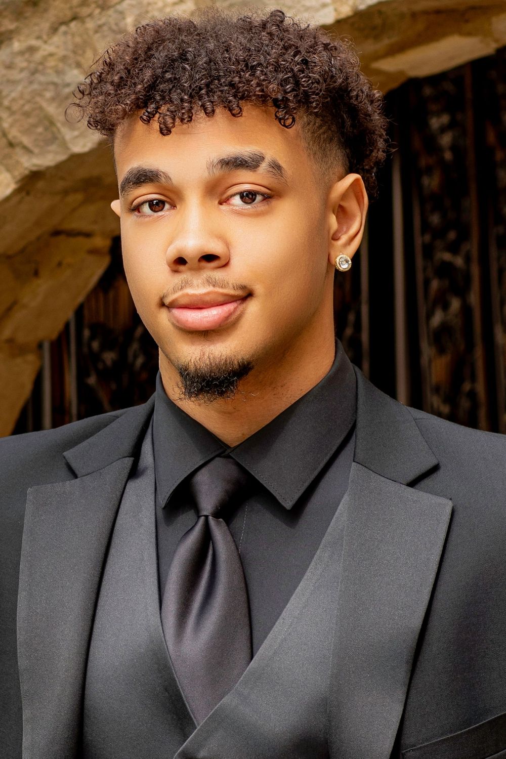 Formal portrait in classic black suit and tie against stone archway background.