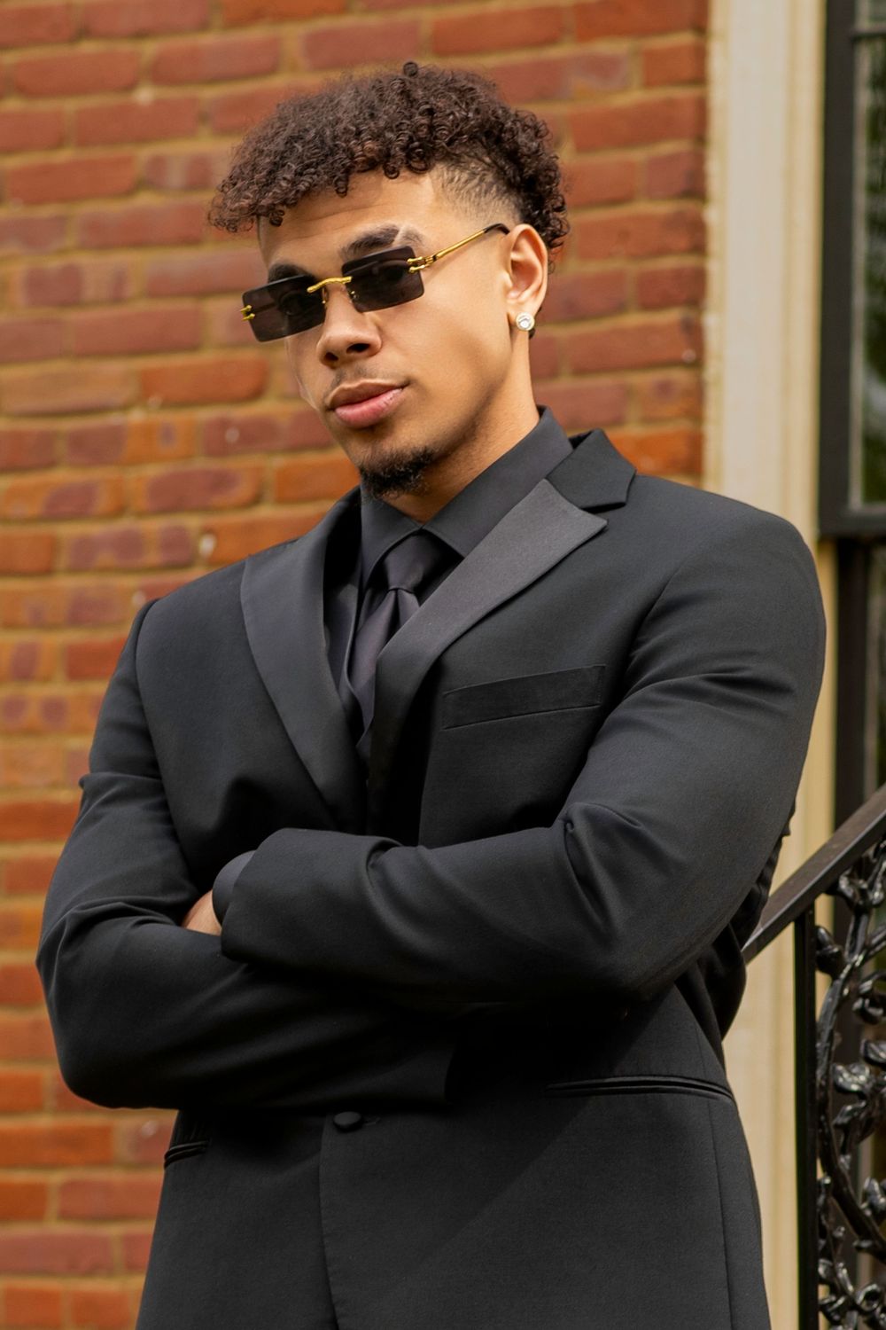 Professional portrait in all black suit and sunglasses against brick wall background.
