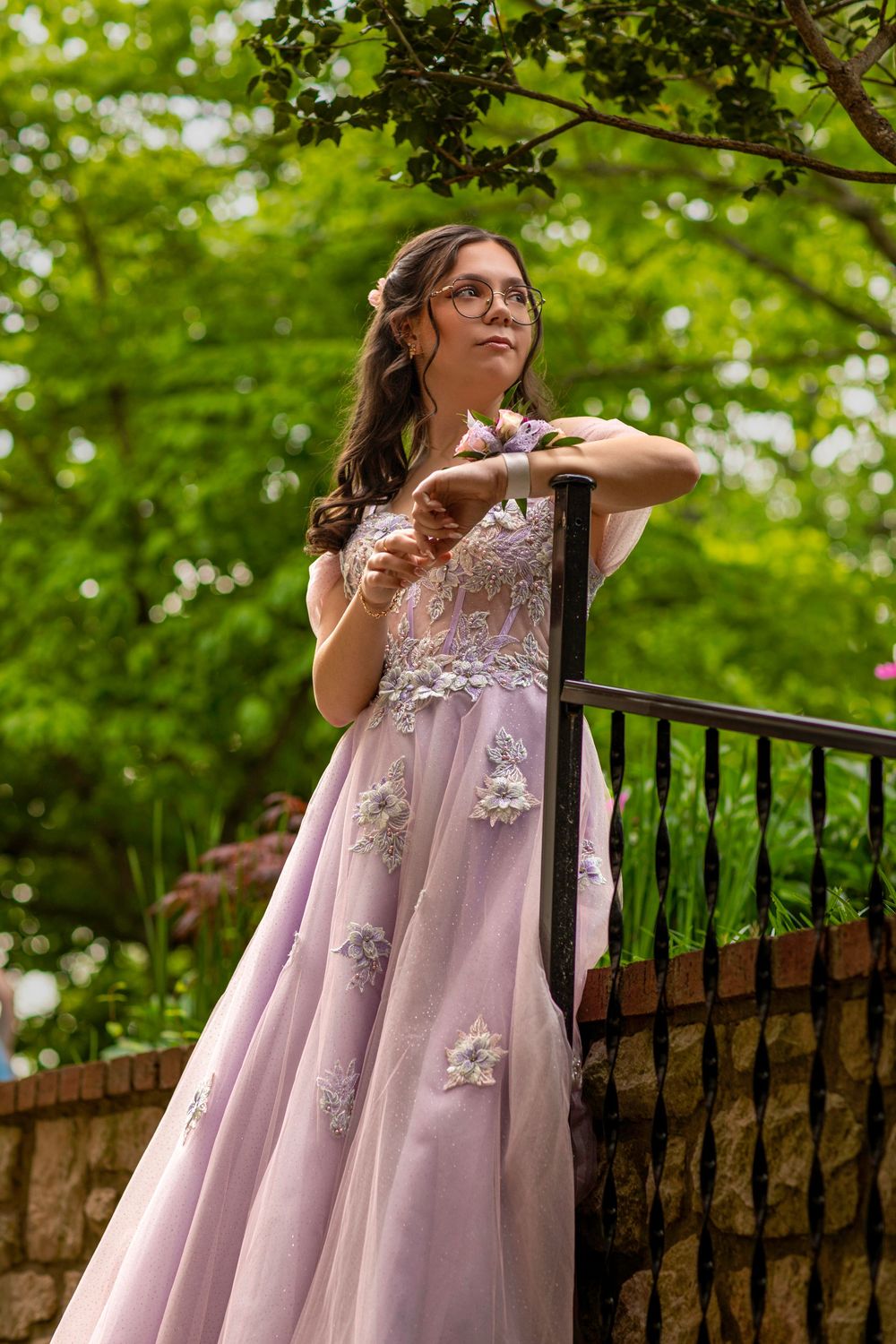 A lavender prom dress with delicate floral appliques photographed against lush green foliage on an outdoor balcony.