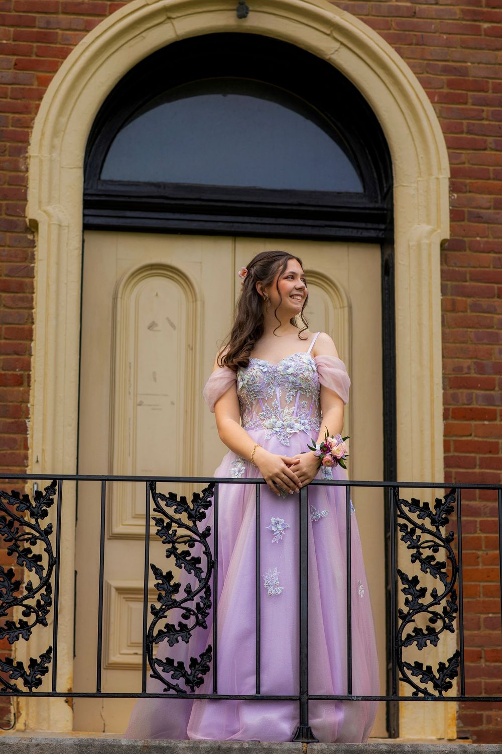 Formal portrait on a brick building balcony featuring a flowing lavender gown with floral details.