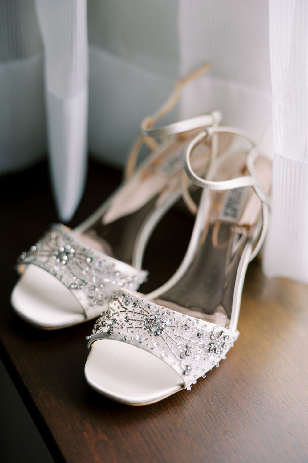 Elegant white bridal sandals with crystal embellishments and ankle straps sit on a wooden surface near sheer curtains at Hall of Springs.