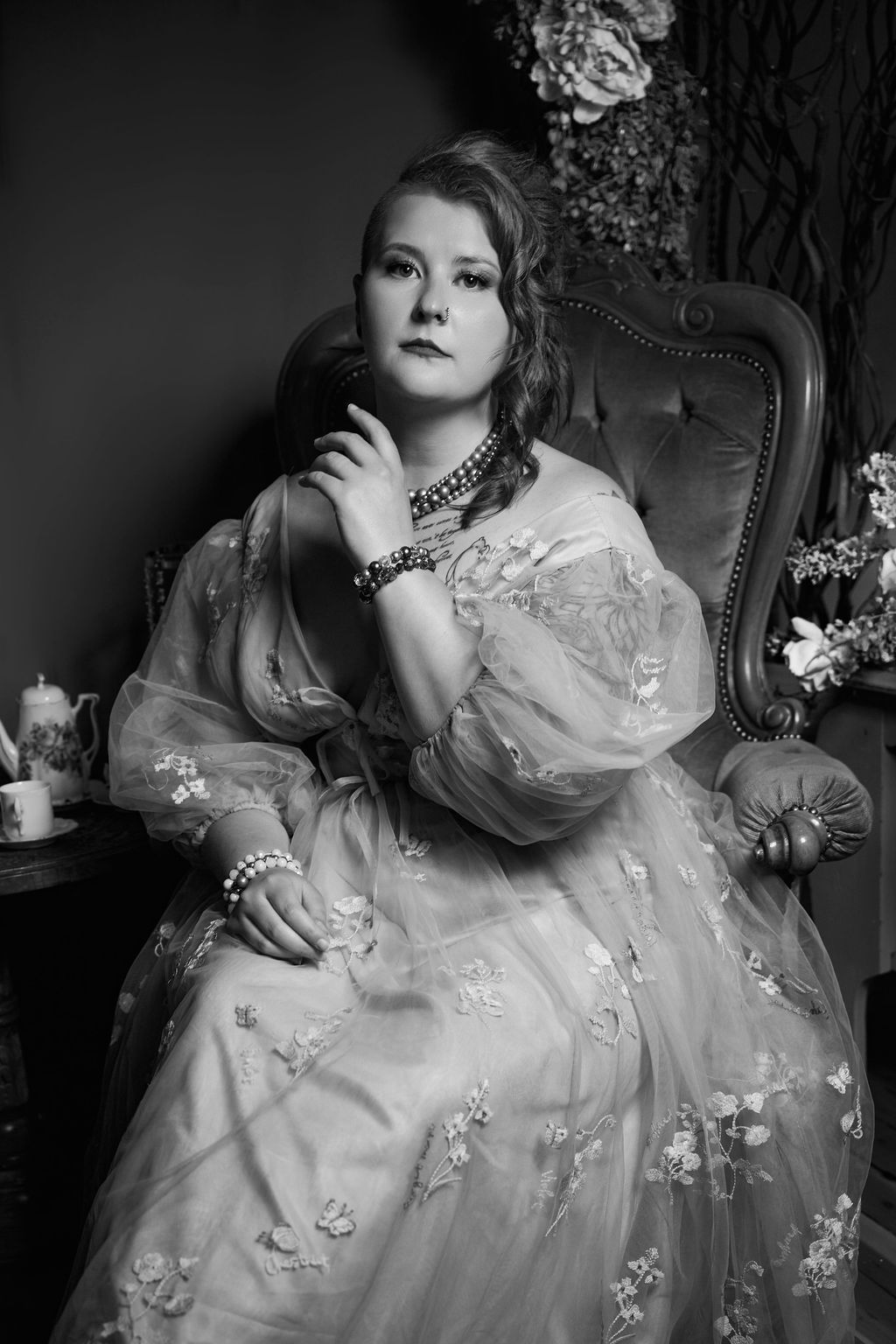 A dramatic black and white Victorian-style portrait featuring an elegant dress with delicate floral embellishments and jewelry.