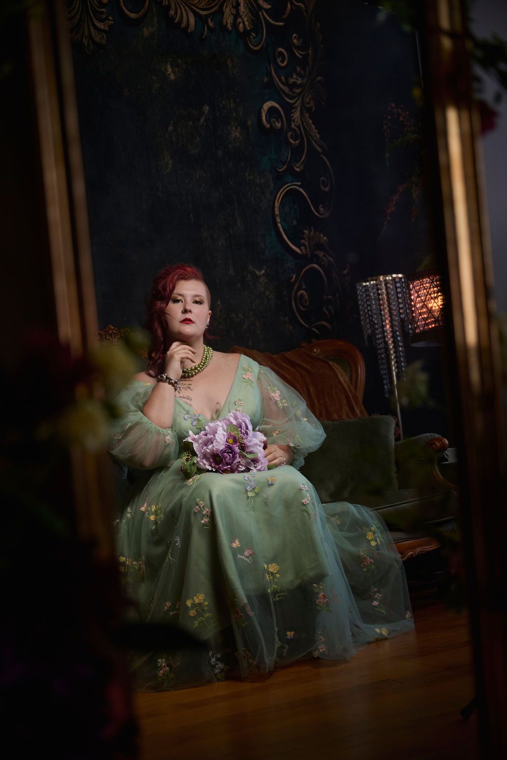 A figure in a mint green vintage gown sits elegantly on a velvet couch holding purple flowers in a moody dark setting.