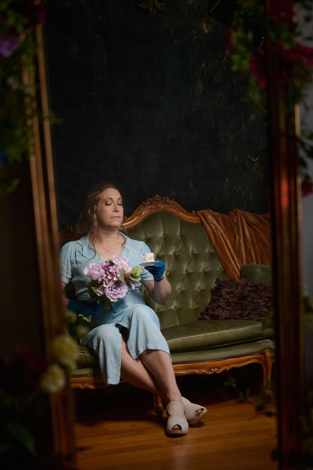 A relaxed scene showing someone in a blue dress resting on an ornate vintage green velvet sofa with flowers.