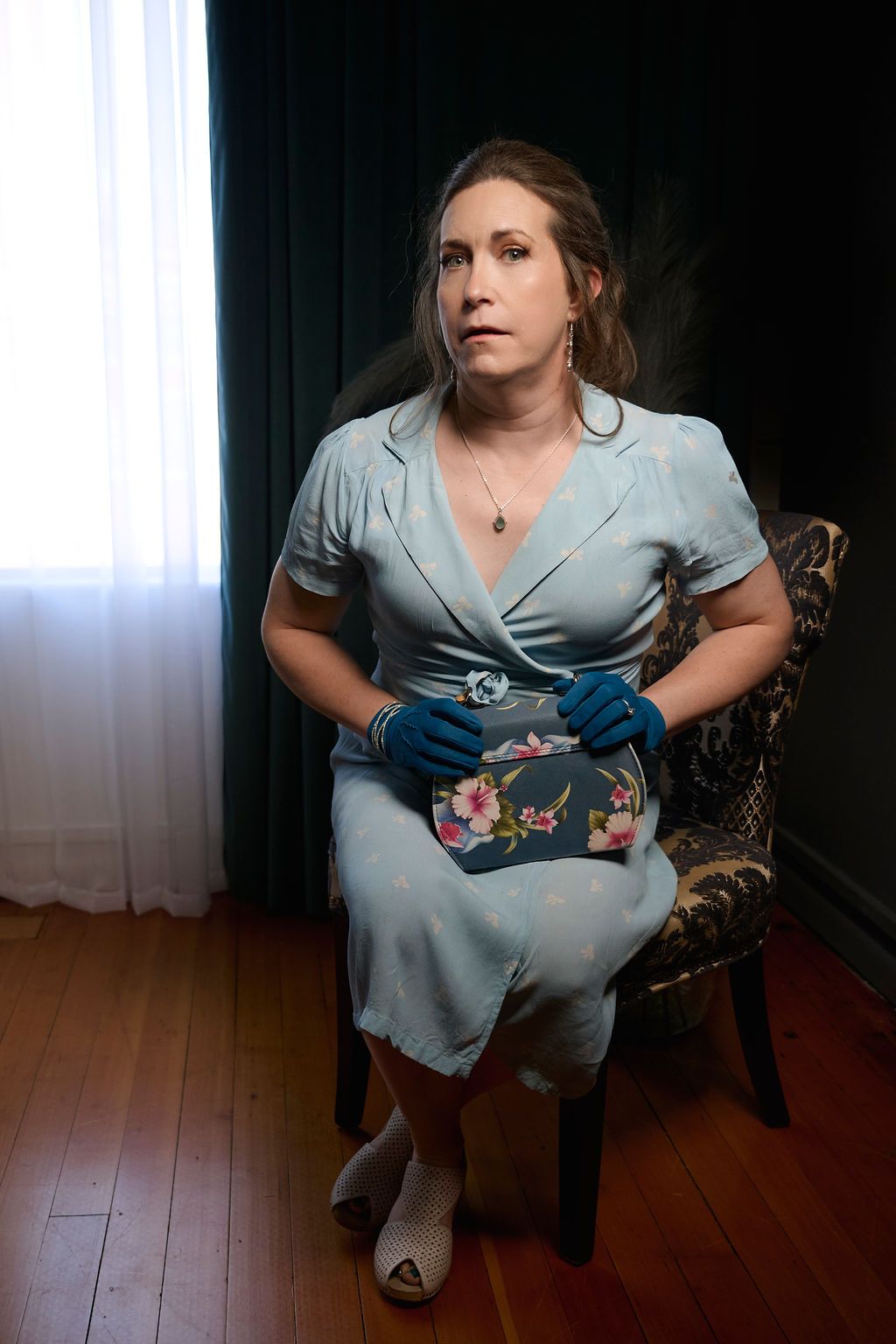 A figure in a light blue vintage-style dress poses on a wooden chair while holding a floral embroidered handbag.