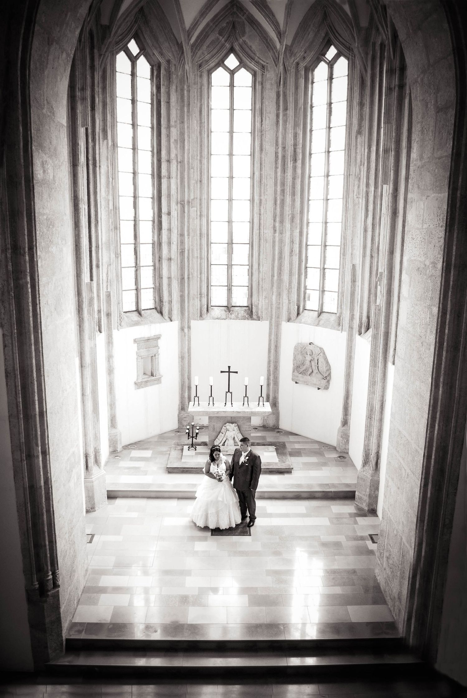 Black and white photograph inside a Gothic cathedral with tall windows and a couple standing at a white altar.