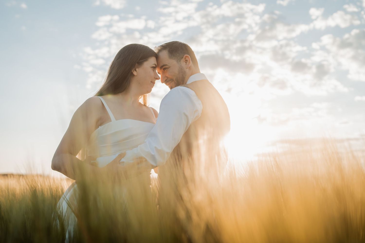 Romantic couple embraces in golden wheat field at sunset with dramatic backlighting through clouds.