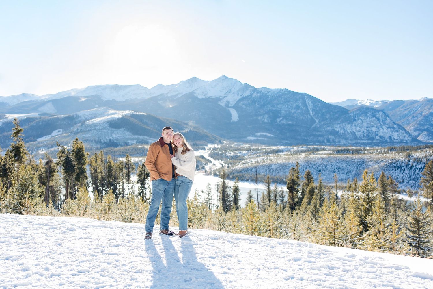 Two people walking through a snowy mountain landscape with scenic mountain peaks in the background.