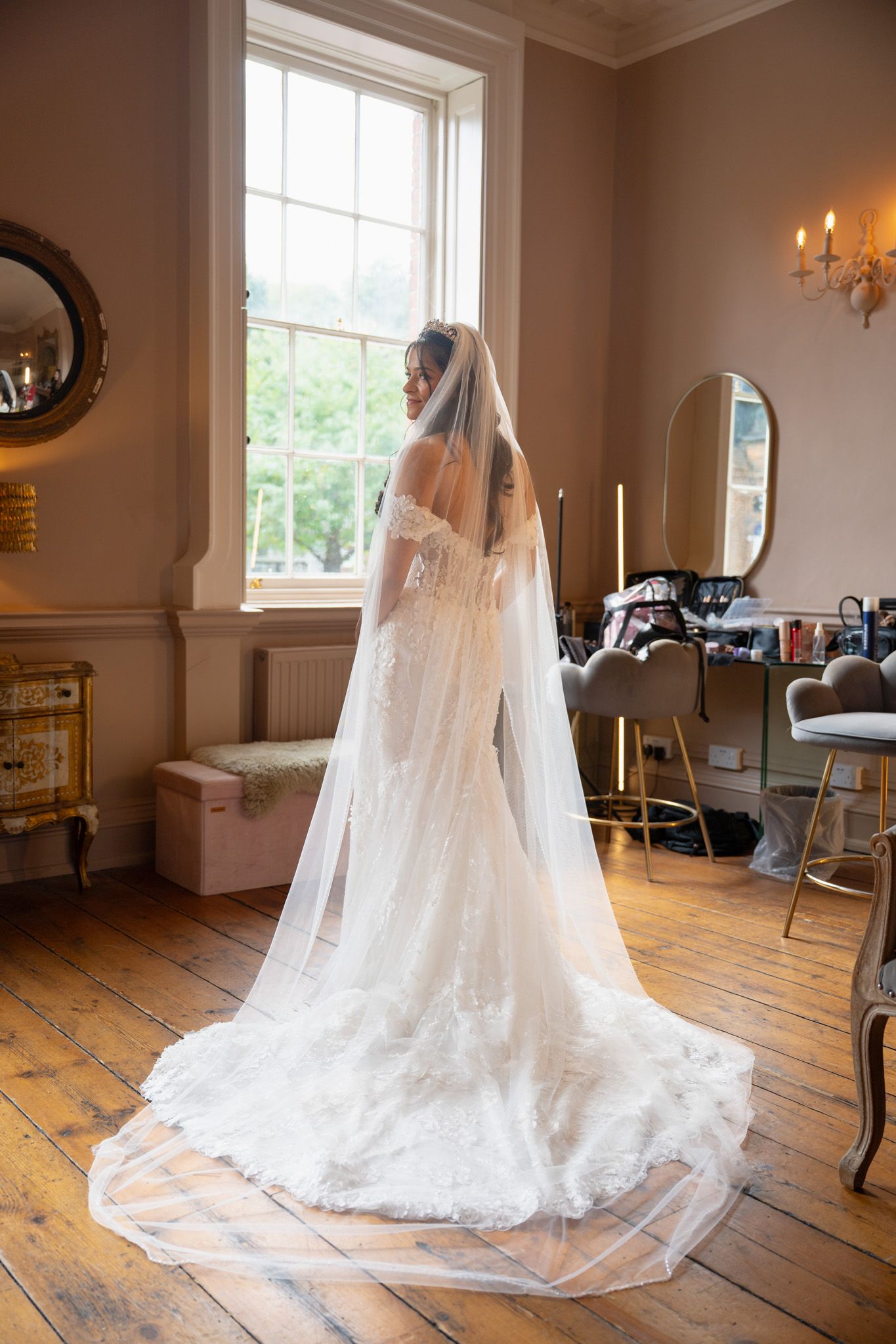 A flowing white wedding dress with a long cathedral veil is displayed in a sunlit room with wooden floors.