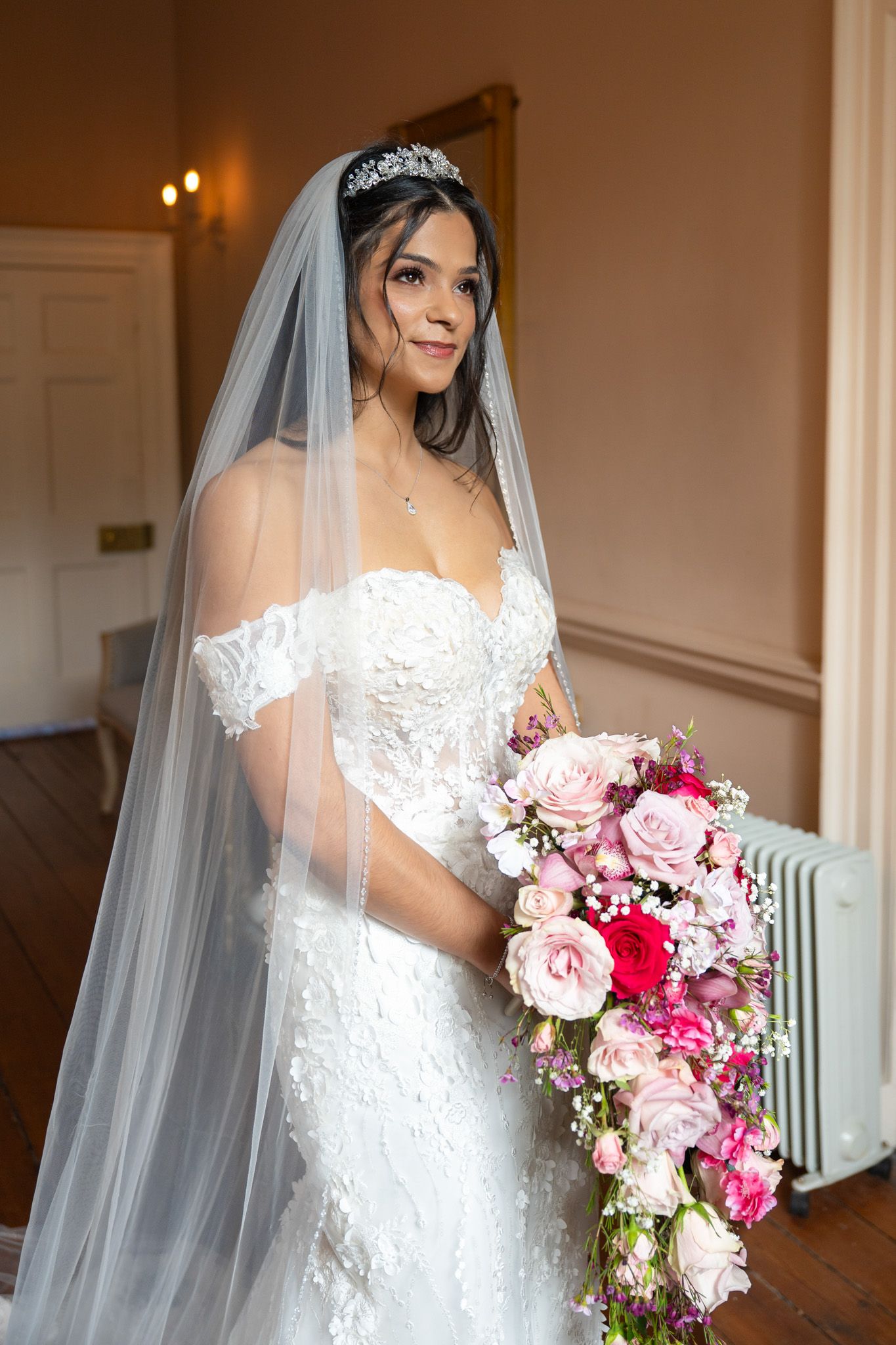 A bride in a white lace wedding dress holds a beautiful pink and red bouquet in an elegant indoor setting.
